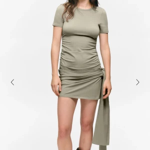 Beige kort klänning med knyt från Monki - Superfin beige kort klänning från Monki med korta ärmar och cool knyt-detalj i sidan. Klänningen har en figurnära passform och är gjord i mjuk bomull, perfekt för en trendig och avslappnad look.