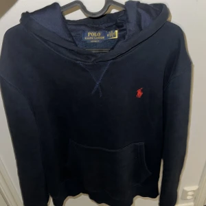 Mörkblå hoodie från Polo Ralph Lauren - Snygg mörkblå hoodie från Polo Ralph Lauren med klassisk röd logga broderad på bröstet. Tröjan har huva, känguruficka och långa ärmar. Perfekt för en chill och stilren look.
