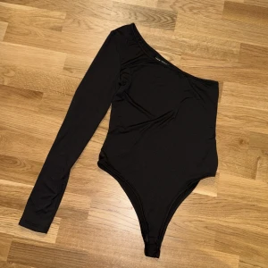 Svart oneshoulder bodysuit från SHEIN - Aldrig använt Svart bodysuit från SHEIN i storlek S med stilren oneshoulder-design och lång ärm. Materialet är mjukt och stretchigt, troligtvis polyester, vilket gör den bekväm och följsam. Perfekt för dig som gillar minimalistisk och trendig stil.