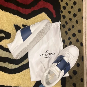 Valentino Garavani Open sneakers vit/blå - Snygga Valentino Garavani Open sneakers i vitt skinn med bred blå läderdetalj över vristen. Klassisk rund tå, vita snören och mönstrad sula. Kommer med original dustbag. Perfekta för dig som vill ha en clean men lyxig look.ingen låda 