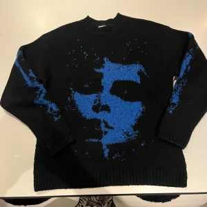 Stickad tröja  - Unik svart stickad tröja från Weekday med ett stort blått ansikte som motiv på framsidan och blå detaljer på ärmarna. 