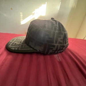 Svart Fendi keps med FF-mönster - Säljer en svart keps från Fendi med ikoniskt FF-mönster över hela kepsen. Kepsen har böjd skärm med svart kant och justerbar rem baktill med Fendi-logga. Materialet är textil med exklusiv känsla och mönstret ger en lyxig streetwear-vibe.