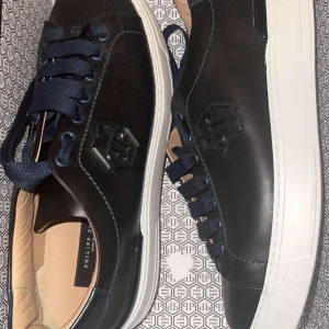 Svarta sneakers från Philipp Plein - Snygga svarta sneakers från Philipp Plein med vit sula och mörkblå snörning. Skorna har en stilren design i skinn med logotyp i 3D på sidan. Perfekta för dig som vill ha en exklusiv och modern look.