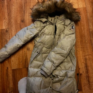 Beige Parajumpers Long Bear dunjacka - Säljer en beige Parajumpers Long Bear Eco dunjacka med fluffig päls på huvan och snyggt emblem på ärmen. Jackan är lång, har dragkedja och praktiska fickor med knappar. Perfekt för kalla vinterdagar och har en riktigt chill vibe. Har fläckar bak (se bild) och hål i luvan (se bild) använt skick därav pris 