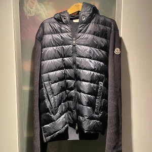 Svart quiltad cardigan Moncler - Svart cardigan från Moncler med quiltad front och huva i glansigt material, samt stickade ärmar med Moncler-logga på vänster arm. Jackan har dragkedja framtill och två sidofickor med dragkedja. Perfekt för dig som gillar street och exklusiv vibe. Snöre tillkommer!