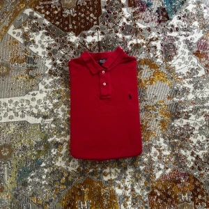 Röd pikétröja från Polo Ralph Lauren i storlek S - Klassisk röd pikétröja från Polo Ralph Lauren med broderad marinblå logga på bröstet. Tröjan har korta ärmar, krage och två knappar framtill. Tillverkad i bomull för en skön och luftig känsla i storlek S
