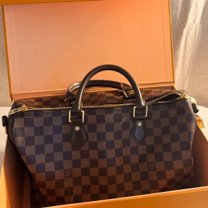 Louis Vuitton Damier handväska i strl 35. Helt ny! - Säljer en klassisk Louis Vuitton Speedy 35 i Damier Ebene-mönster. Väskan är brunrutigt med mörkbruna skinnhandtag, guldfärgade metalldetaljer och dragkedja. Kommer med originalbox, dustbag och lås. Perfekt storlek för vardagens alla prylar.