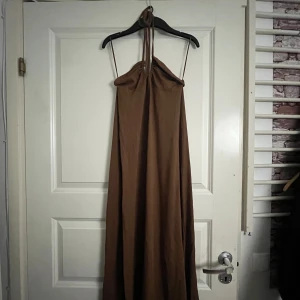 Brun långklänning från Gina Tricot - Säljer en snygg brun långklänning från Gina Tricot i storlek XS. Klänningen har halterneck med smala band och är ärmlös. Den har en enkel, flowy passform och är tillverkad i ett mjukt tyg som känns skönt mot huden. Perfekt för varma dagar och somriga vibes. Aldrig använd då den inte passade mig utan endast testad.