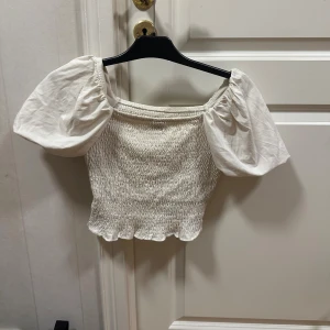 Vit croppad blus från Gina Tricot - Supersöt vit blus från Gina Tricot i storlek S. Modellen är croppad med smockad, ribbad kropp och puffiga korta ärmar. Blusen har en fyrkantig halsringning och volangkant kant nertill.