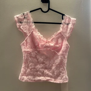 Rosa spetsig linnetopp med rosett - Söt ljusrosa linnetopp i spets med blommigt mönster och liten rosett framtill. Toppen har breda axelband och v-ringning, perfekt för en romantisk look. Croppad modell som ger en trendig vibe. Prislappen är kvar