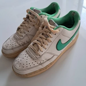 Nike Air Force 1 vit/grön EUR 41 - Nike Air Force 1 sneakers i vitt skinn med gröna detaljer och grön mesh runt ankeln. Klassisk låg modell med perforerad tå, beige snörning och gummisula. Swoosh-logga i grönt på sidorna och blå Nike-tryck på hälen. Perfekt för dig som gillar retrovibbar.