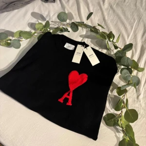 Svart stickad tröja från Ami Paris - Svart stickad tröja från Ami Paris med ikoniskt rött hjärta och bokstaven A broderat på bröstet. Tröjan har rund halsringning och ribbade muddar. Tillverkad i mjuk bomull för en skön känsla och stilren look.