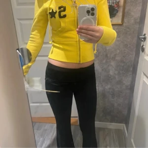 Gul hoodie från HIDDEN CULT - Säljer en gul hoodie från HIDDEN CULT med dragkedja, huva och snörning. Tröjan har svarta detaljer med broderad text och glittriga paljetter framtill. Croppad passform och ribbade muddar. Perfekt för dig som gillar streetstyle och färg!