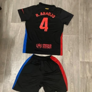 FC Barcelona fotbollströja R. Araujo - Säljer en svart FC Barcelona fotbollströja med R. Araujo och nummer 4 på ryggen. Tröjan har korta ärmar, röda och blå sidopaneler, samt klubbmärke och Spotify-logga framtill. Materialet är lätt och andas, perfekt för match eller träning.