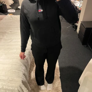 Svart hoodie från Tommy jeans  - Svart hoodie från Tommy jeans med klassisk logga på bröstet. Tröjan har huva med snörning och en stor magficka framtill. Perfekt för dig som gillar en enkel och stilren look med streetvibe.