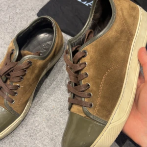 Bruna sneakers från Lanvin i mocka uk 7 - Snygga sneakers från Lanvin i brun mocka med gröna lackdetaljer på tån och runt kanten. Skorna har bruna snören, vit sula och insida i svart skinn. Perfekta för dig som gillar en stilren men unik look.