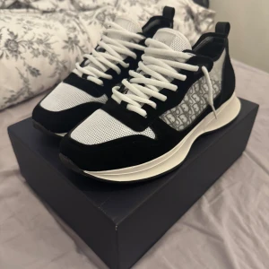 Svarta och vita Dior b25 - Snygga Dior sneakers med svart och vit färg, mesh och mockadetaljer. Sidan har det klassiska Dior-mönstret i grått och vitt. Skorna har rund tå, platt sula och vita snören. Perfekta för dig som vill ha en lyxig och modern look.