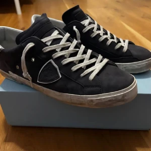 Philippe Model skor - Svarta Philippe Model sneakers i mocka med vit häl och vita snören. Snyggt sköldmärke på sidan och metallnitar som detalj. Platt sula med lite sliten look för extra edge. Perfekta för dig som gillar stilrena och coola sneakers.