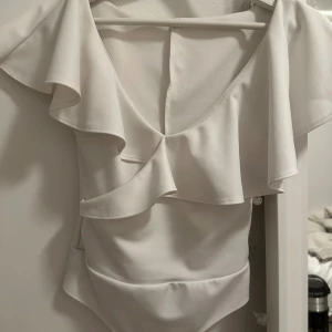 Vit volang body från NLY One XS - Säljer en vit body från NLY One i storlek XS. Toppen har breda volanger över axlarna och en snygg v-ringning framtill. Materialet är mjukt och stretchigt, perfekt för dig som gillar en feminin och trendig look.