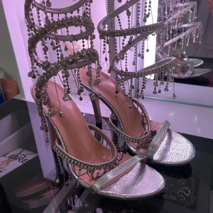 Glittriga silver sandaler med smyckade band - Unika sandaler i metallic silver med hög klack och öppen tå. Skorna har flera spiralformade band upp längs benet, dekorerade med gnistrande kedjor och hängande pärlor för extra bling. Ytan har ett lätt ormmönster som ger en lyxig känsla.