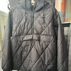 Svart quiltad pufferjacka med huva - Quiltad svart pufferjacka med huva och snörning, half zip framtill och en stor ficka över magen. Jackan har långärmade ärmar och en loose passform, perfekt för kalla dagar. Snyggt rutmönster över hela jackan ger en cool vibe.