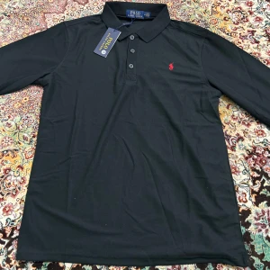 Svart långärmad pikétröja Polo Ralph Lauren - Svart långärmad pikétröja från Polo Ralph Lauren med klassisk krage och tre knappar framtill. Tröjan har det ikoniska röda Polo-logot broderat på bröstet och är gjord i mjuk bomull som känns skön mot huden. Perfekt för dig som gillar stilren och enkel design.