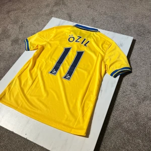 Arsenal Özil 11 gul bortatröja Nike - Gul Arsenal bortatröja med blå detaljer, Özil 11-tryck på ryggen. Klassisk pikékrage i blått med gula ränder, broderat Arsenal-märke och Nike-logga på bröstet. Tillverkad i lätt och ventilerande polyester, perfekt för match eller samling.