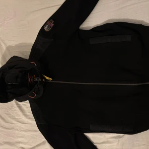 Parajumper Cardigan  - Svart vindjacka från Parajumpers med huva och snygga orange detaljer. Jackan har dragkedja framtill, patch på ärmen och diskreta fickor. Materialmix med polyester och stickad känsla på vissa partier. Perfekt för dig som gillar stilren och sportig look. Storlek M, passar även S vilket jag har.