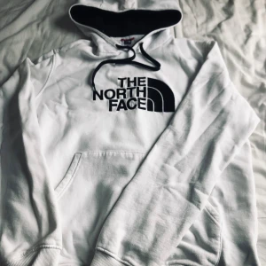 Vit hoodie från The North Face  - Säljer en vit hoodie från The North Face i storlek L. Tröjan har svart logotyp på bröstet, svart snörning i huvan och mjukt foder på insidan. Klassisk känguruficka framtill och ribbade muddar vid ärmslut och nederkant. Perfekt för chill dagar.