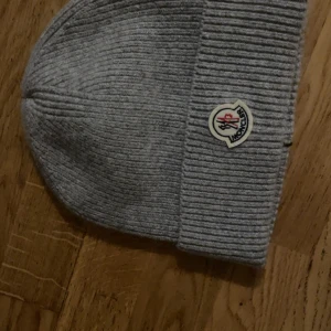 Grå ribbstickad mössa från Moncler - Snygg grå ribbstickad mössa från Moncler med klassisk Moncler-logga framtill. Mössan är tillverkad i mjukt material och har en uppvikt kant för extra stilpoäng. Perfekt accessoar för kyliga dagar och en clean look.
