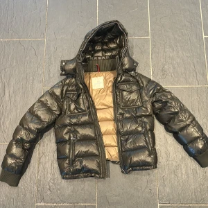 Moncler jacka. Pris kommer höjas i vinter, billigast nu - Mörk grön dunjacka från Moncler med avtagbar huva och ribbade muddar. Jackan har två bröstfickor med lock, två sidofickor med dragkedja och beige insida. Klassisk Moncler-logga på ärmen. Mycket fint skicka men skavanker finns, men inget som påverkar jackan fina hel het. 