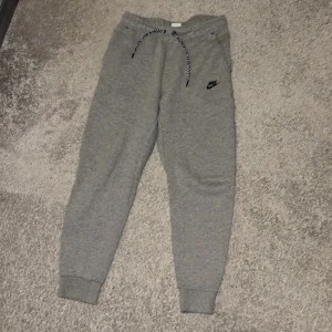 Grå mjukisbyxor från Nike, storlek M - Snygga grå mjukisbyxor från Nike med svart logga på vänster ben. Byxorna har ribbade muddar vid bensluten, praktisk snörning i midjan och fickor med dragkedja på baksidan. Perfekta för chill eller träning, tillverkade i mjukt bomullsmaterial.