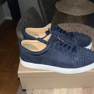Mörkblå Louboutin sneakers med nitar - Säljer ett par mörkblå sneakers från Christian Louboutin i mocka med coola ton-i-ton nitar på tån. Skorna har vita platta sulor, rund tå och matchande blå snörning. Insidan är beige och de har en lyxig känsla som sticker ut. Mycket bra skick bara att 2 spikar har åkt av där framme. Men syns knappt!