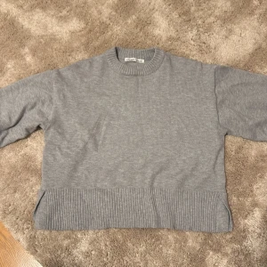 Grå stickad tröja från Pull&Bear - Säljer en grå stickad tröja från Pull&Bear i storlek XS. Tröjan har en rund halsringning, ribbade muddar vid hals, ärmslut och nederkant samt slitsar på sidorna. Perfekt för dig som gillar enkel och clean stil.