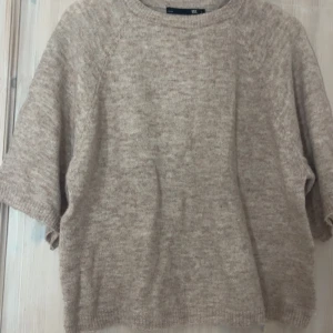 Beige stickad tröja från 157 - Mysig beige stickad tröja från 157 med rund hals och korta ärmar. Tröjan har en enkel, loose passform och är perfekt för lager-på-lager. Materialet är mjukt och har en diskret melerad look. Passar dig som gillar chill och stilren vibe.