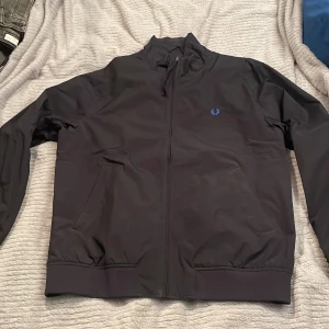 Marinblå Fred Perry bomberjacka L - Säljer en marinblå bomberjacka från Fred Perry i storlek L. Jackan har klassisk ribbad krage, muddar och nederkant, samt två sidofickor. Materialet är syntet och jackan har en blå broderad Fred Perry-logga på bröstet. Perfekt för dig som gillar stilren och sportig look.