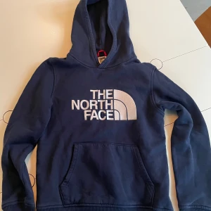 Marinblå hoodie från The North Face - Snygg marinblå hoodie från The North Face med stor vit logga på bröstet. Tröjan har huva, magficka och ribbade muddar vid ärmslut och nederkant. Perfekt för dig som gillar streetwear och vill ha en bekväm och stilren look.
