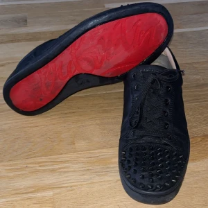 Svarta Louboutin sneakers med nitar - Säljer ett par svarta sneakers från Christian Louboutin med klassisk röd sula och coola svarta nitar på tån. Skorna har snörning och är tillverkade i skinn, vilket ger en lyxig känsla. Perfekta för dig som vill sticka ut med en edgy och exklusiv stil. Pris går att diskutera. Passar även storlek 43