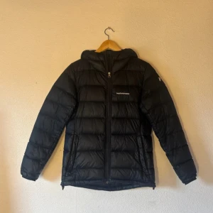 Svart dunjacka Peak Performance S - Svart dunjacka från Peak Performance i storlek S. Jackan har huva, dragkedja framtill och diskret logga på bröstet och ärmen. Supervarm och skön, sparsamt använd! Har en liten defekt (se bild) men inget som syns vid användning!! Kan skickas snabbt.😊