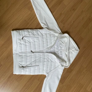 Vit quiltad pufferjacka med huva - Snygg vit pufferjacka med huva och dragkedja framtill. Jackan har quiltade partier på kroppen och släta ärmar, samt två fickor med dragkedja. Perfekt för dig som gillar en clean och sportig look.👀 ALDRIG använd!!! Så den är ny 👍 hör av er för fler frågor!!!  Står ej för frakt 