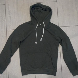 Mörkgrön hoodie från Pier One - Säljer en stilren mörkgrön hoodie från Pier One med vita snören och klassisk känguruficka. Tröjan har lång ärm och huva, perfekt för chill dagar. Materialet är mjukt och skönt, troligtvis bomullsmix. Enkel och snygg design som funkar till det mesta.