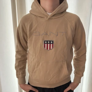 Beige hoodie från GANT  - Snygg beige hoodie från GANT med broderat logotyp och ett stort emblem med stjärnor och ränder på bröstet. Tröjan har huva, magficka och långa ärmar. Storlek 146-152, 11-12 år. 