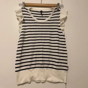 Söt topp - Säljer en söt vit topp med svarta horisontella ränder från Laura Torelli. Toppen har volangdetaljer vid axlarna och är ärmlös med rund halsringning. Materialet känns mjukt och stretchigt, perfekt för dig som gillar en chill och fräsch stil.