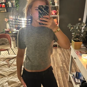 Grå croppad stickad tröja - Säljer en grå croppad stickad tröja med korta ärmar och rund hals. Tröjan har en mjuk och skön känsla, perfekt för dig som gillar en enkel men snygg look. Passar dig som vill ha en lite kortare modell till jeans eller kjol.