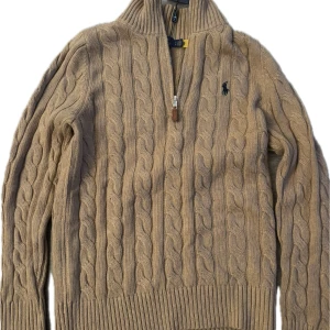 Beige kabelstickad tröja Polo Ralph Lauren - Snygg beige kabelstickad tröja från Polo Ralph Lauren med half zip och hög krage. Tröjan har långärmad passform, ribbade muddar och klassisk broderad logga på bröstet. Perfekt för dig som gillar stilrena och tidlösa plagg.