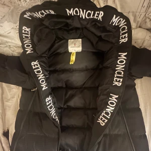 Svart Moncler dunjacka med huva - Säljer en svart dunjacka från Moncler med stor huva och tydlig logga tryckt längs hela huvkanten. Jackan har dragkedja, vadderad design och klassisk Moncler-patch på ärmen. Storlek L