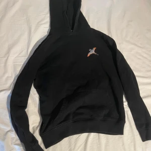 Svart hoodie Axel Arigato med broderad fågel - Svart hoodie från Axel Arigato med broderad fågel i grått och orange på bröstet. Tröjan har huva, magficka och långa ärmar. Mjuk bomullskvalitet och klassisk passform, perfekt för chill dagar. Snygg detalj med fågeln som ger extra stil.