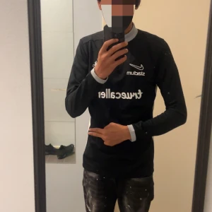 Svart AIK långärmad Nike fotbollströja - AIK fotbollströja från Nike med lång ärm, svart bas och vita detaljer vid krage och sidor. Stadium och Truecaller-loggor på bröstet samt AIK:s klubbmärke. Materialet är lätt och andas, med meshpaneler på sidorna för extra ventilation. Halv dragkedja framtill.