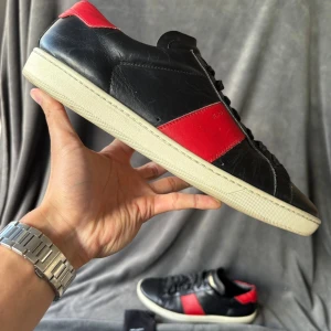 Saint Laurent svarta och röda sneakers - Snygga sneakers från Saint Laurent i svart skinn med röda detaljer på sidan och hälen. Klassisk rund tå, vit platt sula och svart snörning. Märket är tryckt i guld på plösen och sula. Perfekt för dig som gillar stilrena och lyxiga sneakers.
