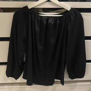 Svart blus med knyt från 157 XS - Svart blus från 157 i storlek XS med rundad hals och knyt framtill. Blusen har långärmade puffiga ärmar och är gjord i ett lätt, krinklat tyg som ger en cool struktur. Perfekt för dig som gillar enkel men snygg stil.